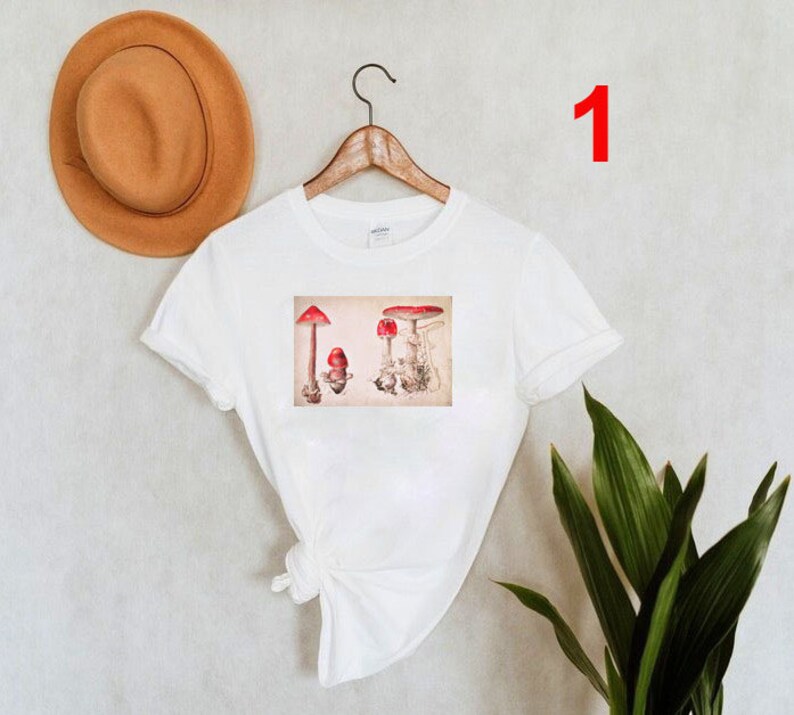 May include: White t-shirt with a vintage illustration of red mushrooms on the front. The illustration is in a rectangular frame.