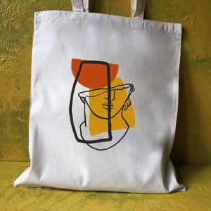 Op de afbeelding: Een witte tote bag met een zwarte lijntekening van een buste, gedeeltelijk bedekt door een gele en oranje geometrische vorm. De buste is omlijnd met een zwarte lijn en heeft een eenvoudig, gestileerd gezicht.