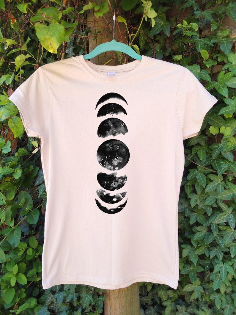 May include: A light beige t-shirt featuring a black watercolor design of the phases of the moon. The graphic shows the moon's cycle from crescent to full and back. The shirt is hanging on a light blue hanger against a backdrop of green foliage.