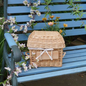 May include: A wicker casket with a white ribbon and a wooden nameplate sits on a blue wooden bench. The bench is surrounded by white flowers and green foliage.
