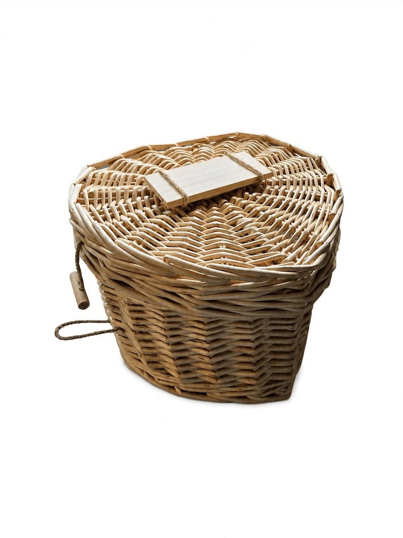 May include: A heart-shaped woven wicker basket with a lid. The basket features a light tan natural finish and a small rectangular wooden tag attached to the top of the lid with a thin piece of twine. A small wooden toggle is attached to the side.