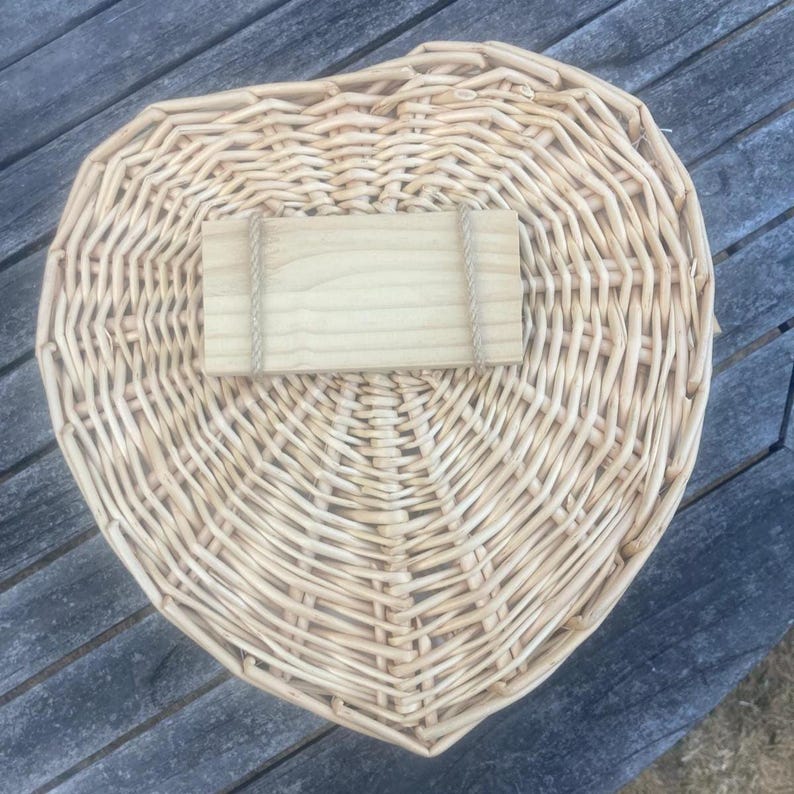 May include: A heart-shaped wicker basket made of light tan woven reeds. Centered on the top of the basket is a rectangular, light-colored wooden plaque attached with thin, natural-toned twine wrapped around the ends of the wood piece.