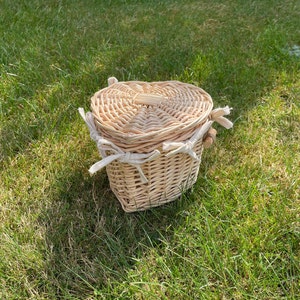 May include: A small, heart-shaped wicker basket with a white fabric lining and a wooden handle. The basket is sitting on a bed of green grass.