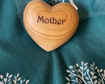 Pode incluir: Um ornamento de madeira em forma de coração com a palavra "Mãe" gravada. O ornamento está sobre um tecido verde com um desenho de árvore prateada.