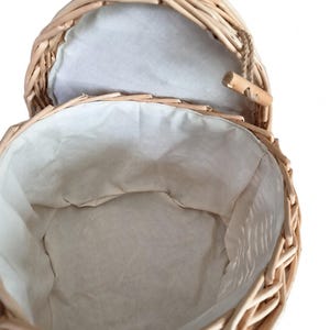 May include: A round woven wicker basket with a light tan natural finish. The interior is lined with a plain off-white fabric. The basket features a hinged lid attached with a small wooden toggle closure and a short piece of brown twine.