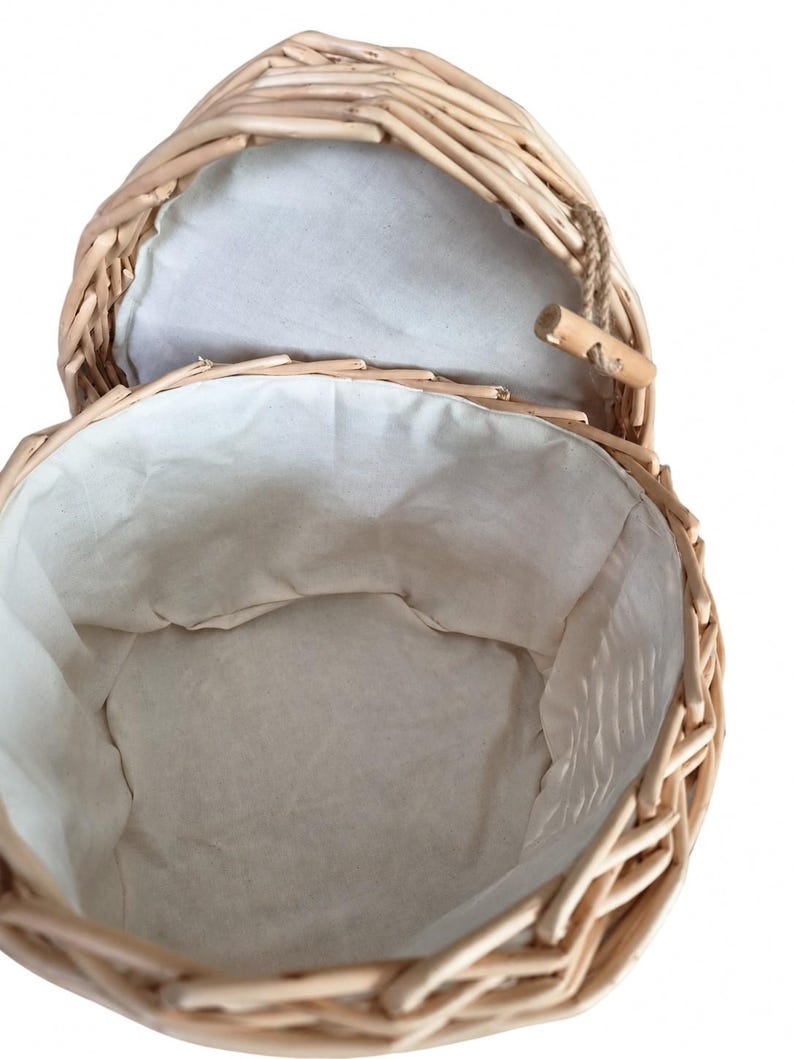 May include: A round woven wicker basket with a light tan natural finish. The interior is lined with a plain off-white fabric. The basket features a hinged lid attached with a small wooden toggle closure and a short piece of brown twine.