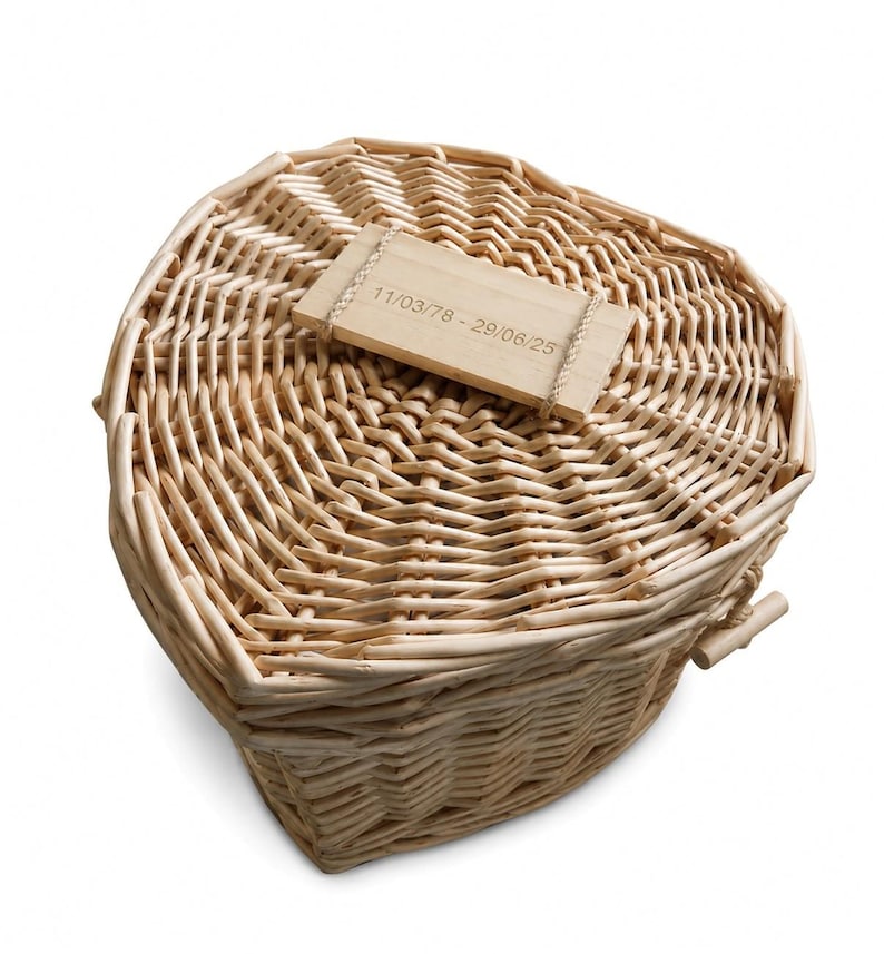 May include: A heart-shaped wicker basket with a light tan natural finish. A rectangular wooden plaque is attached to the lid with thin twine, featuring the engraved text 11/03/78 - 29/06/25. The basket has a woven texture and a lid.