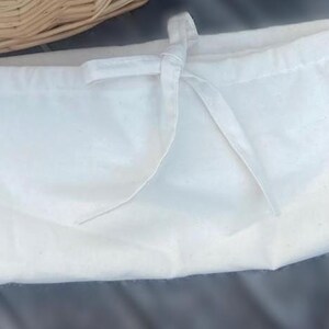 May include: A rectangular off-white fabric drawstring pouch featuring a simple tie closure at the top center. The material has a subtle speckled texture throughout, and the bag is constructed with clean, straight stitched seams along the edges.