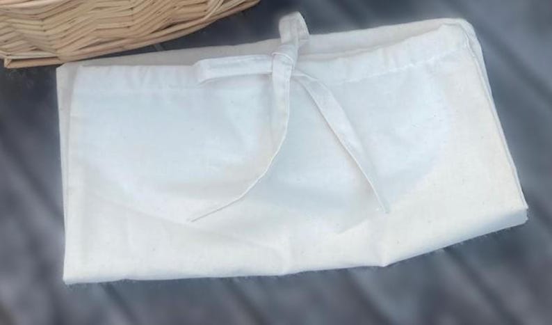 May include: A rectangular off-white fabric drawstring pouch featuring a simple tie closure at the top center. The material has a subtle speckled texture throughout, and the bag is constructed with clean, straight stitched seams along the edges.