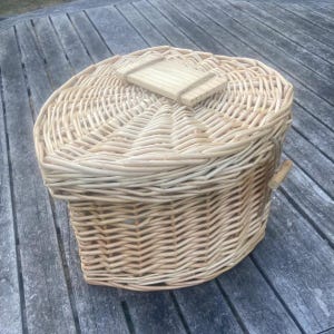 May include: A light tan woven wicker basket with a heart-shaped design and a matching lid. The lid features a small, rectangular wooden handle centered on top. The basket has a sturdy, textured weave pattern throughout its entire exterior structure.