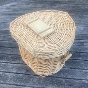 May include: A light tan woven wicker basket with a rounded, heart-like shape and a matching lid. The lid features a small, rectangular wooden handle centered on top, and the basket includes a small wooden toggle closure on the side.