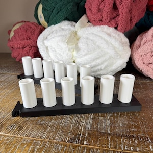 May include: Two black rectangular bases each featuring six upright white cylindrical holders arranged in a single row. The holders are smooth and hollow at the top, designed to support yarn spools or similar items, positioned in front of several large skeins of yarn.
