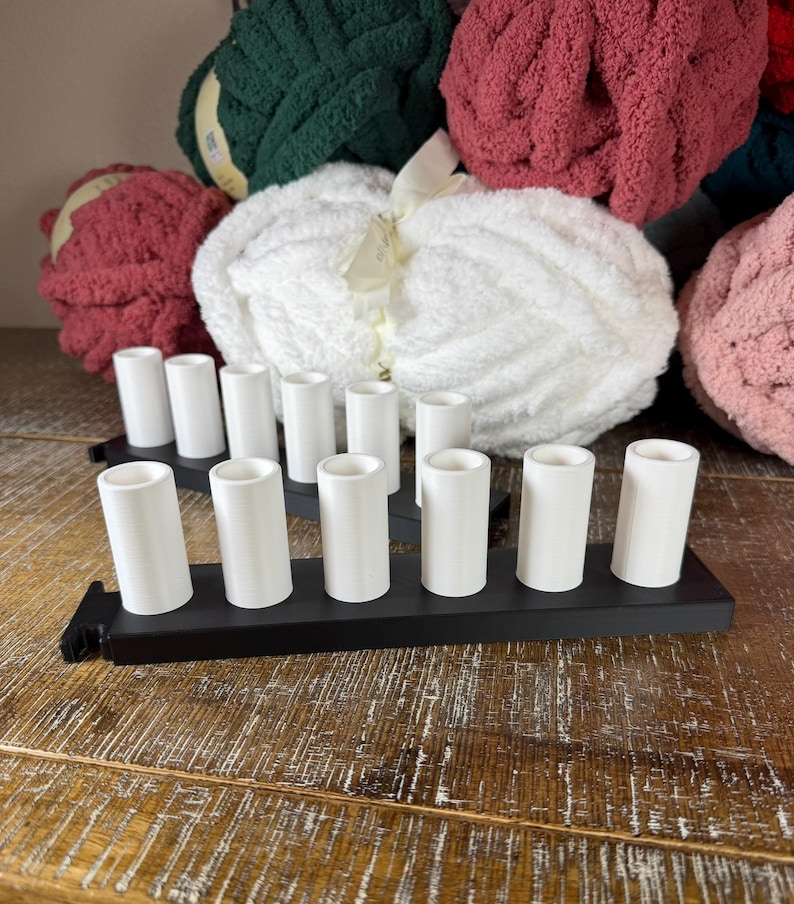 May include: Two black rectangular bases each featuring six upright white cylindrical holders arranged in a single row. The holders are smooth and hollow at the top, designed to support yarn spools or similar items, positioned in front of several large skeins of yarn.