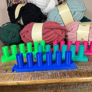 May include: A collection of rectangular plastic yarn spool holders in bright blue, green, and pink, each featuring five vertical cylindrical pegs. Behind the holders are several skeins of chunky, soft-textured yarn in white, black, teal, red, and pink.