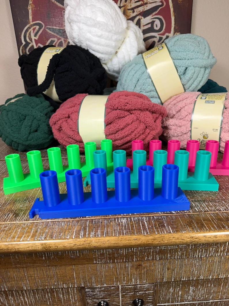 May include: A collection of rectangular plastic yarn spool holders in bright blue, green, and pink, each featuring five vertical cylindrical pegs. Behind the holders are several skeins of chunky, soft-textured yarn in white, black, teal, red, and pink.