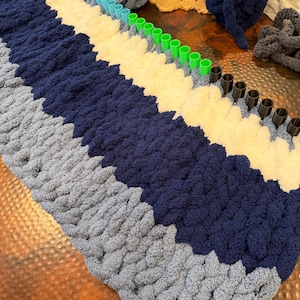 May include: A rectangular knitting loom with rows of red, light blue, green, and black pegs. The loom is partially filled with thick, chunky yarn in alternating stripes of light blue, dark navy blue, and cream white, with extra yarn balls nearby.