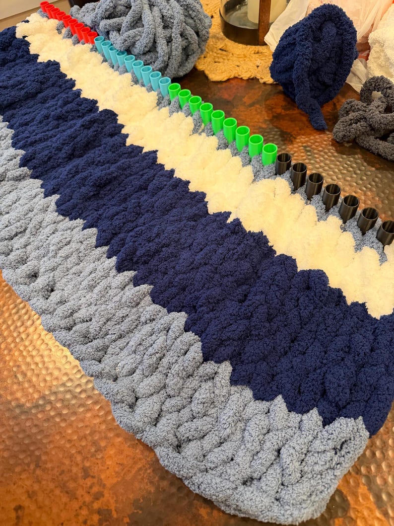 May include: A rectangular knitting loom with rows of red, light blue, green, and black pegs. The loom is partially filled with thick, chunky yarn in alternating stripes of light blue, dark navy blue, and cream white, with extra yarn balls nearby.