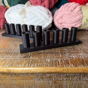 May include: Two black rectangular organizers featuring six vertical cylindrical holders each. These matte black plastic stands are designed with uniform circular openings arranged in a single row, providing a structured base for holding various small items.