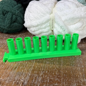 May include: A bright green plastic knitting loom tool featuring a rectangular base with nine evenly spaced vertical pegs. The tool is positioned in front of several skeins of chunky yarn in white, dark green, and black, with the brand name Mellobello visible on the white yarn label.