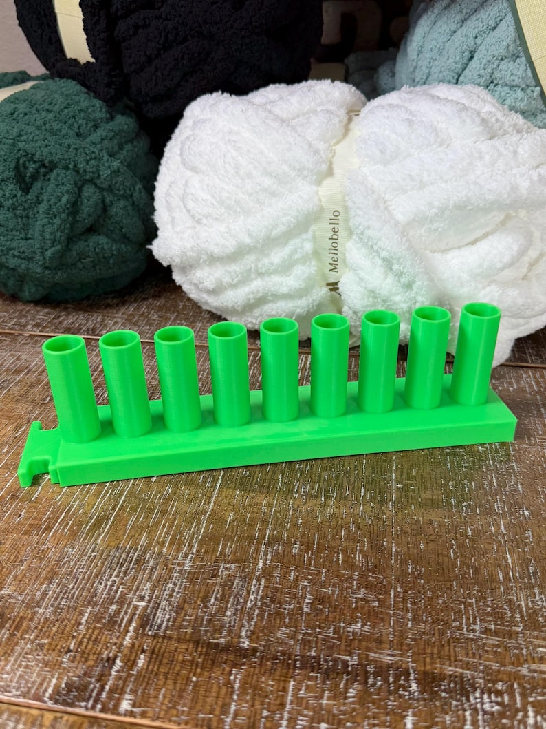 May include: A bright green plastic knitting loom tool featuring a rectangular base with nine evenly spaced vertical pegs. The tool is positioned in front of several skeins of chunky yarn in white, dark green, and black, with the brand name Mellobello visible on the white yarn label.