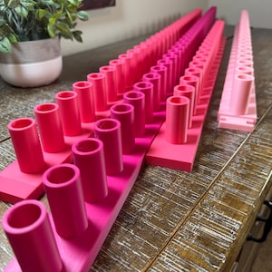 May include: Three long, rectangular pink plastic organizers featuring multiple vertical cylindrical pegs arranged in a single row. Each organizer has a flat base and uniform hollow tubes extending upward, designed for holding small items in place.
