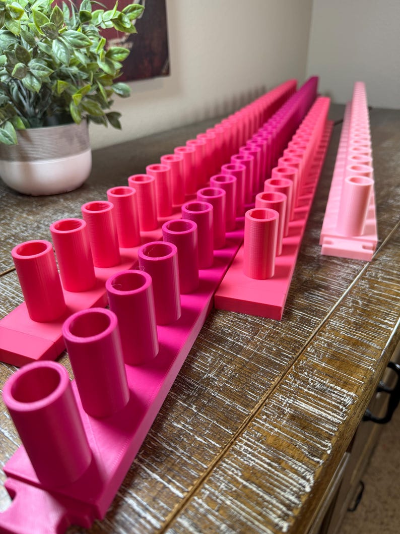 May include: Three long, rectangular pink plastic organizers featuring multiple vertical cylindrical pegs arranged in a single row. Each organizer has a flat base and uniform hollow tubes extending upward, designed for holding small items in place.