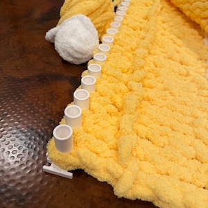 May include: A white plastic knitting loom with a row of pegs holding a thick, yellow, chunky yarn blanket in progress. Beside the loom sits a ball of yellow yarn and a smaller ball of white yarn, both featuring a soft, plush, textured finish.