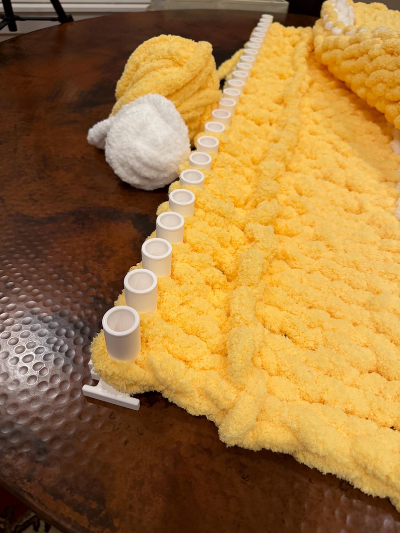 May include: A white plastic knitting loom with a row of pegs holding a thick, yellow, chunky yarn blanket in progress. Beside the loom sits a ball of yellow yarn and a smaller ball of white yarn, both featuring a soft, plush, textured finish.