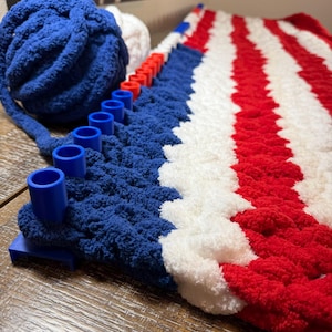 May include: A blue, white, and red striped blanket in progress on a blue plastic knitting loom. A large ball of blue yarn sits next to the loom, with a smaller ball of white yarn partially visible behind it, showing the textured yarn stitches.