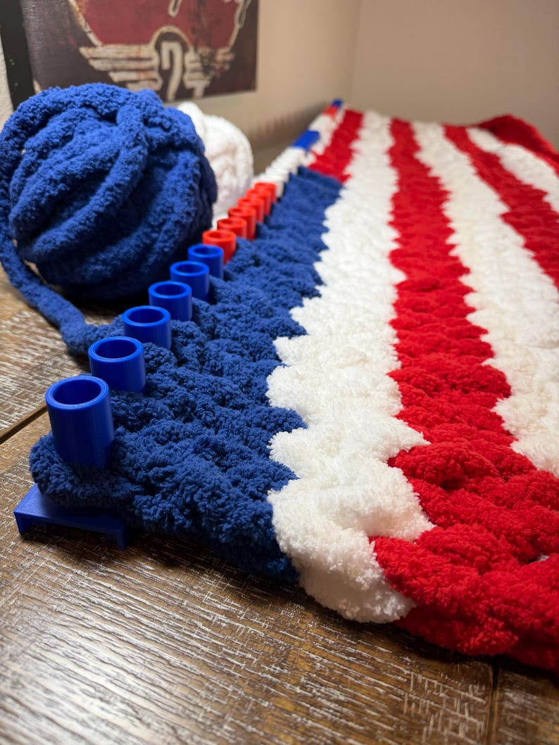 May include: A blue, white, and red striped blanket in progress on a blue plastic knitting loom. A large ball of blue yarn sits next to the loom, with a smaller ball of white yarn partially visible behind it, showing the textured yarn stitches.