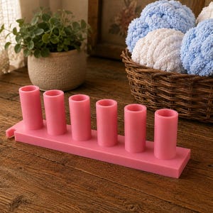 May include: A pink plastic organizer stand featuring six vertical cylindrical holders arranged in a single row on a rectangular base. The object is smooth, solid in color, and designed with a small tab extension on one side of the base structure.