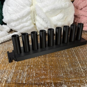 May include: A matte black organizer stand featuring nine vertical cylindrical holders arranged in a single row on a rectangular base. The stand is constructed with a textured finish and includes a small notched cutout on the left side of the base.