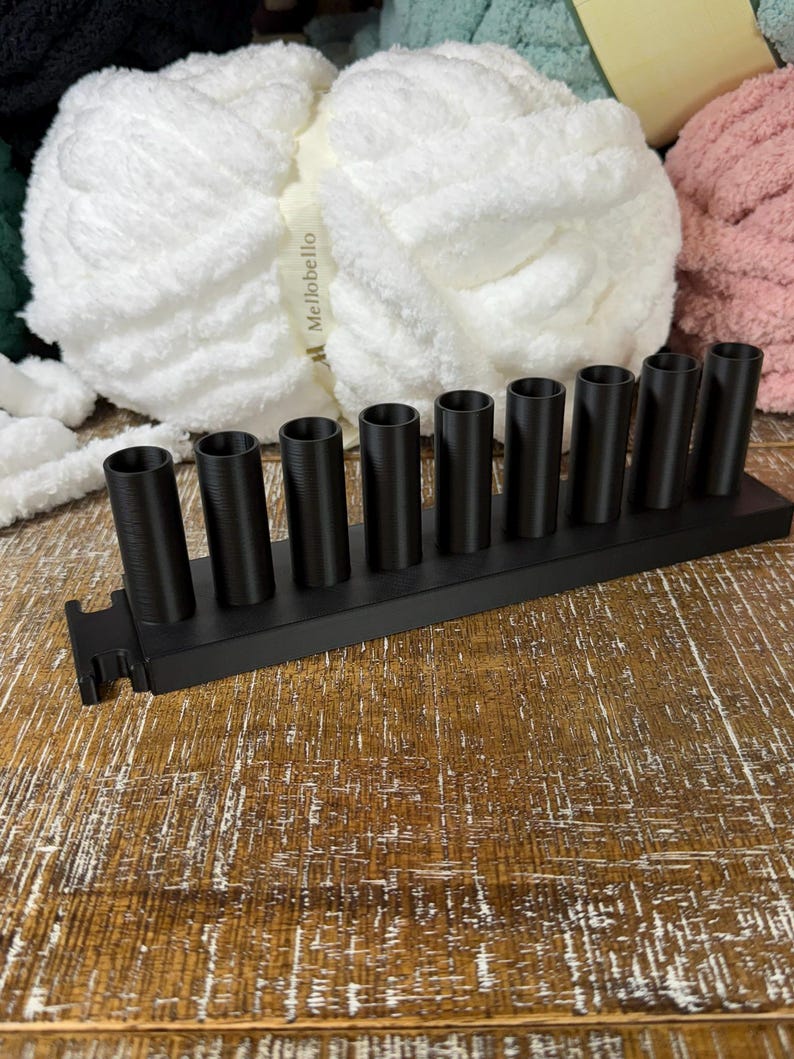 May include: A matte black organizer stand featuring nine vertical cylindrical holders arranged in a single row on a rectangular base. The stand is constructed with a textured finish and includes a small notched cutout on the left side of the base.