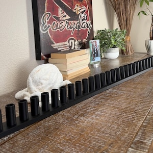May include: A long, narrow, matte black candle holder base featuring a continuous row of numerous cylindrical sockets designed to hold multiple taper candles in a straight, linear arrangement across a flat surface.