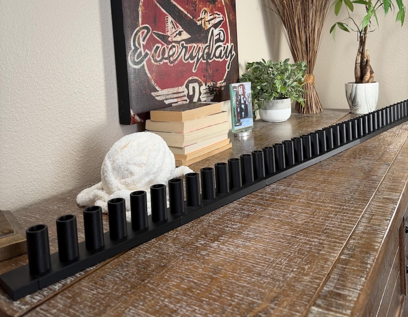May include: A long, narrow, matte black candle holder base featuring a continuous row of numerous cylindrical sockets designed to hold multiple taper candles in a straight, linear arrangement across a flat surface.