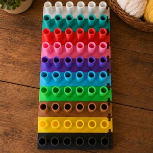 May include: A multi-colored knitting loom set consisting of ten rectangular plastic segments, each featuring six vertical pegs. The segments are arranged in a vertical stack, displaying a spectrum of colors including white, teal, red, pink, purple, blue, green, brown, yellow, and black.