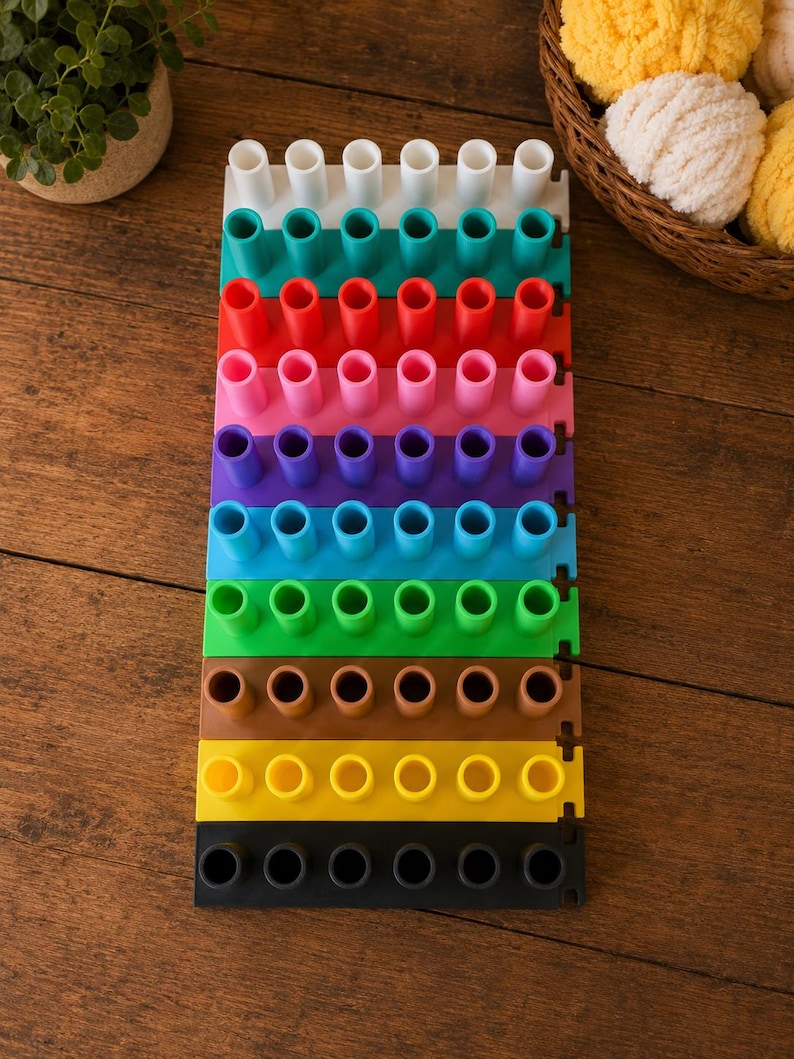 May include: A multi-colored knitting loom set consisting of ten rectangular plastic segments, each featuring six vertical pegs. The segments are arranged in a vertical stack, displaying a spectrum of colors including white, teal, red, pink, purple, blue, green, brown, yellow, and black.