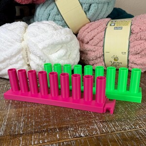 May include: Two rectangular plastic yarn spool holders, one in bright pink and one in neon green, each featuring a row of eight vertical cylindrical pegs. The holders are positioned in front of three large skeins of soft, textured yarn in white, pink, and teal.