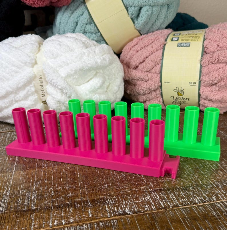May include: Two rectangular plastic yarn spool holders, one in bright pink and one in neon green, each featuring a row of eight vertical cylindrical pegs. The holders are positioned in front of three large skeins of soft, textured yarn in white, pink, and teal.