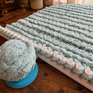 May include: A light blue chunky yarn blanket in progress on a white rectangular knitting loom with evenly spaced pegs. A matching ball of soft, textured light blue yarn sits on a circular blue base next to the loom, showing the material used.