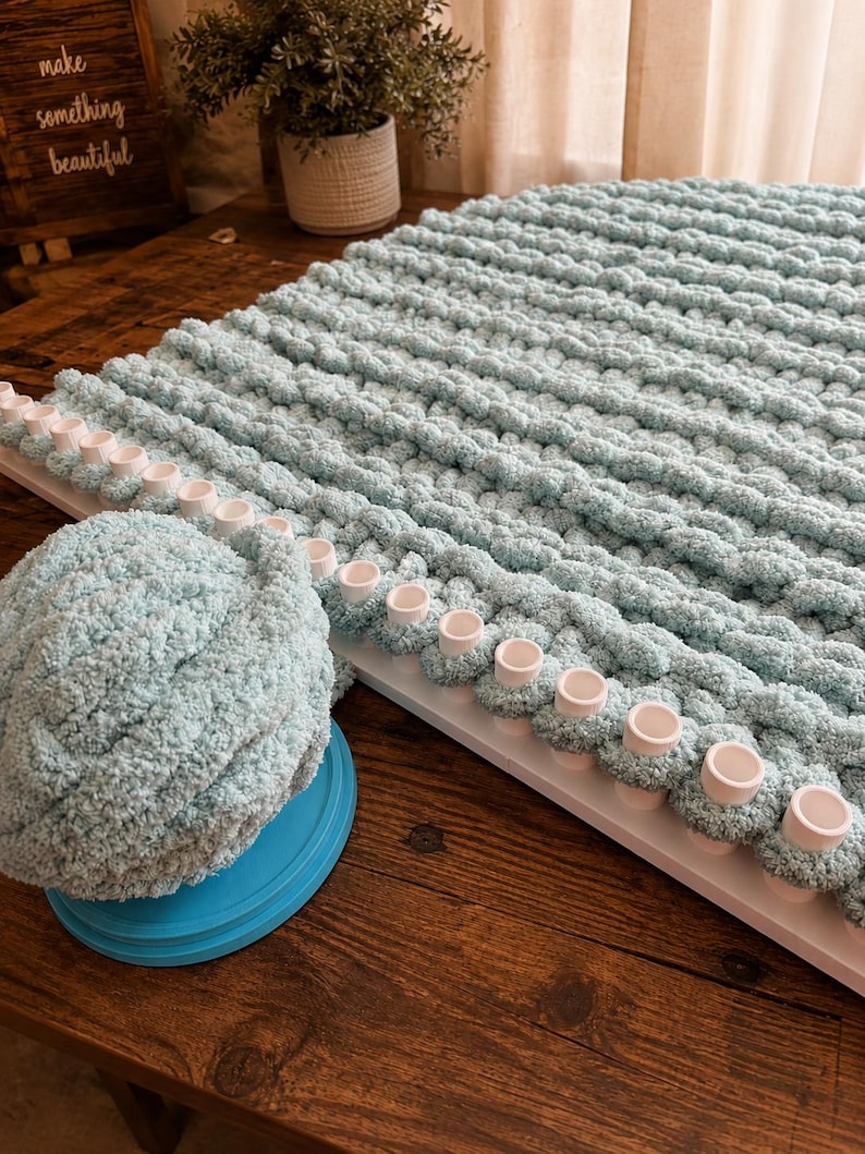 May include: A light blue chunky yarn blanket in progress on a white rectangular knitting loom with evenly spaced pegs. A matching ball of soft, textured light blue yarn sits on a circular blue base next to the loom, showing the material used.