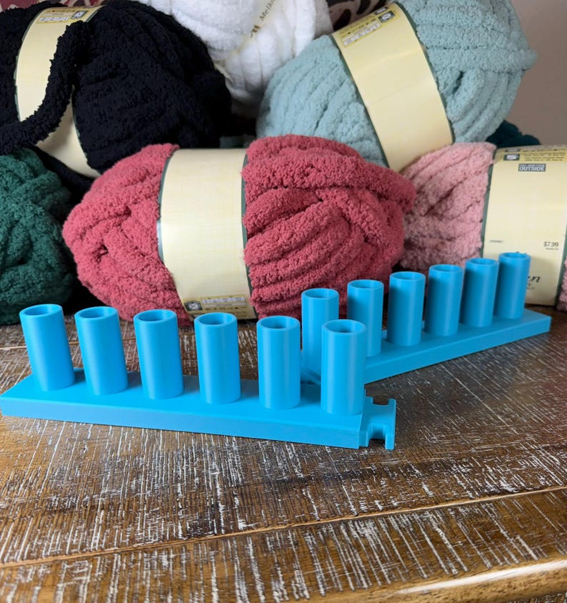 May include: Two bright blue plastic knitting looms with multiple upright pegs, positioned in front of several skeins of chunky yarn in black, white, sage green, dusty rose, and dark green, each wrapped with a tan paper label.