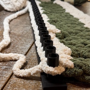 May include: A black plastic knitting loom with a single row of pegs, featuring thick, textured cream-colored yarn looped around the pegs and a section of dark olive green yarn worked into the fabric, with additional yarn skeins nearby.