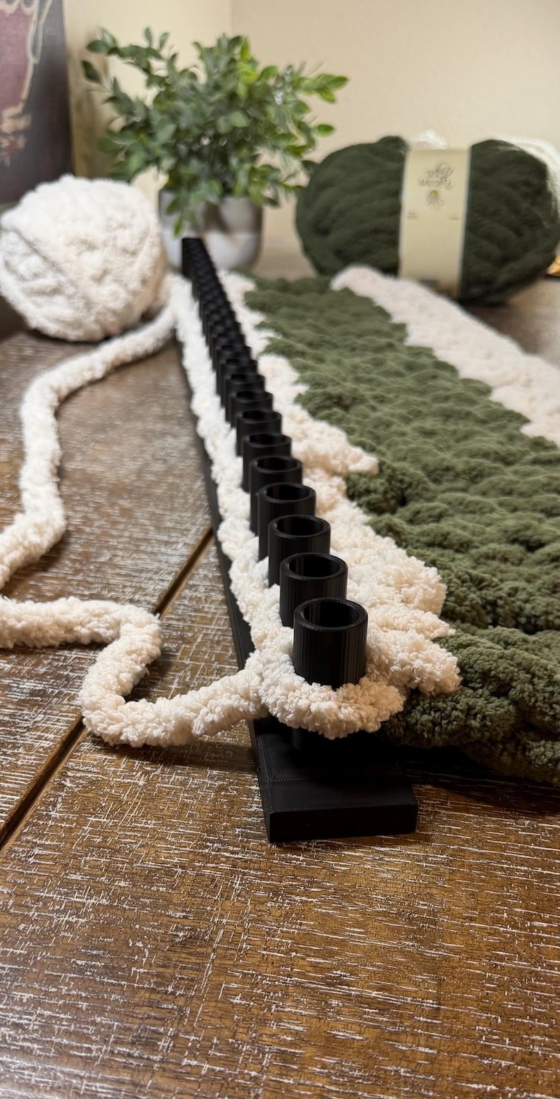 May include: A black plastic knitting loom with a single row of pegs, featuring thick, textured cream-colored yarn looped around the pegs and a section of dark olive green yarn worked into the fabric, with additional yarn skeins nearby.
