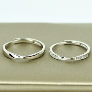 May include: Two silver-colored, adjustable rings. One ring has a smooth, twisted design. The other ring has a similar twisted design, but is embellished with small, clear gemstones. Both rings are displayed on a neutral-toned surface.
