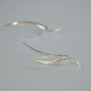 May include: A pair of silver ear cuffs with a wavy design. The cuffs are made of a textured metal and have a delicate, minimalist look.