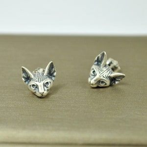 May include: A pair of silver earrings featuring a detailed, three-dimensional design of a Sphynx cat's head.