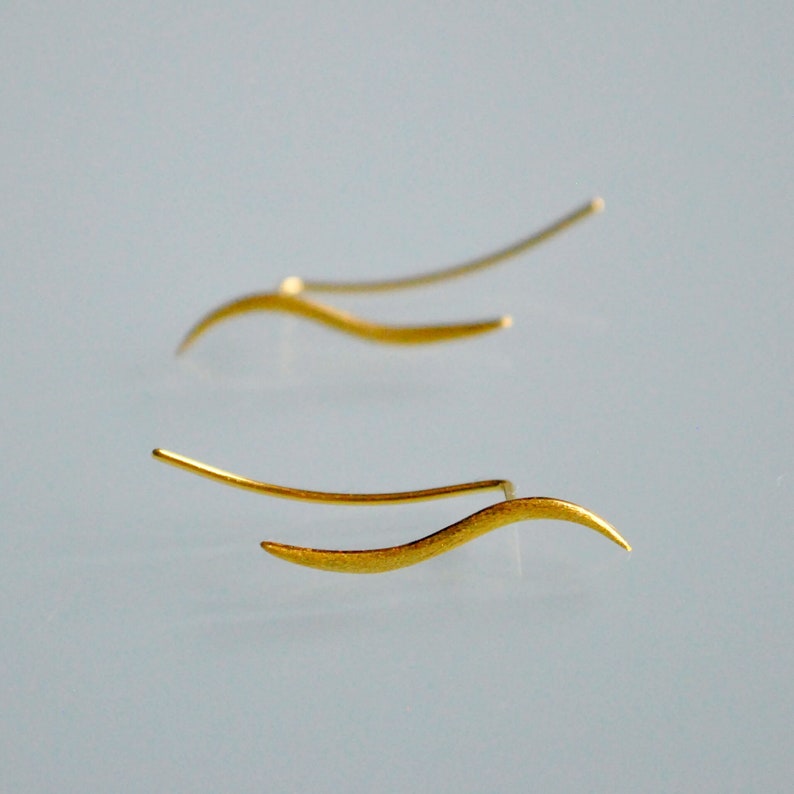 May include: A pair of gold-toned earrings with a wavy design. The earrings are made of thin metal and have a delicate, minimalist look.