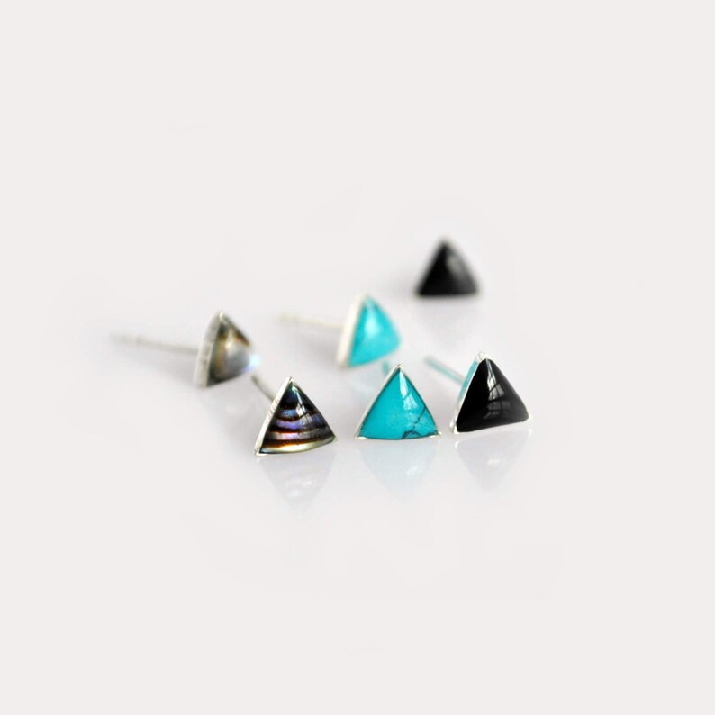 May include: Three silver triangle stud earrings with different colored stones. The earrings feature a turquoise stone, a black stone, and a multi-colored abalone shell.
