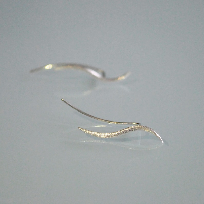 May include: A pair of silver ear cuffs with a wavy design. The cuffs are made of a textured metal and have a delicate, minimalist look.
